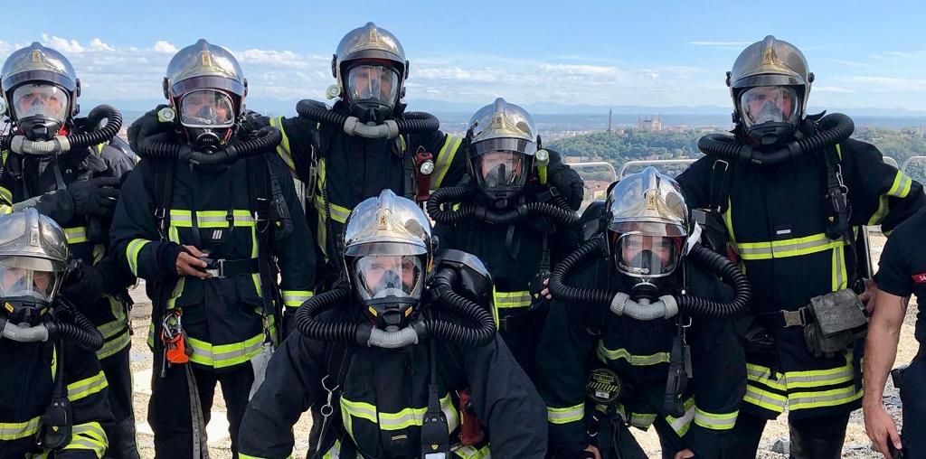 Un groupe de pompiers portant un équipement de protection complet et des casques se tiennent ensemble à l'extérieur par une journée ensoleillée, avec un paysage pittoresque visible en arrière-plan. Devenir Pompier