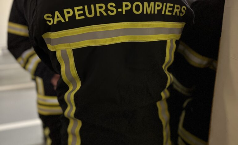 Une personne portant un uniforme de pompier noir avec des bandes réfléchissantes jaunes et les mots « SAPEURS-POMPIERS » dans le dos se tient parmi d'autres personnes habillées de la même manière à l'intérieur. Devenir Pompier